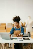 Startup small business SME, Entrepreneur owner African woman using smartphone or tablet taking receive and checking online purchase shopping order to preparing pack product box. in office photo