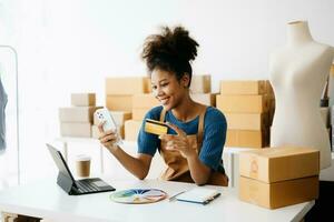 Startup small business SME, Entrepreneur owner African woman using smartphone or tablet taking receive and checking online purchase shopping order to preparing pack product box. in office photo