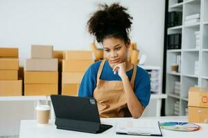 puesta en marcha pequeño negocio pequeño, empresario propietario africano mujer utilizando teléfono inteligente o tableta tomando recibir y comprobación en línea compra compras orden a preparando paquete producto caja. en oficina foto