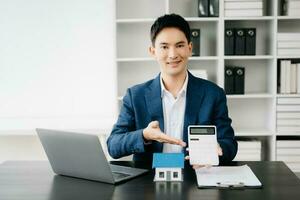 Young real estate agent worker working with laptop and tablet at table in modern office and small house beside it. photo