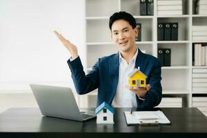 Young real estate agent worker working with laptop and tablet at table in modern office and small house beside it. photo