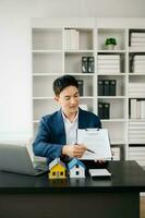 Young real estate agent worker working with laptop and tablet at table in modern office and small house beside it. photo