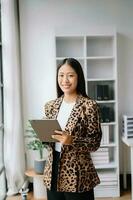 joven atractivo asiático hembra oficina trabajador negocio trajes sonriente a cámara con trabajando bloc, tableta y ordenador portátil documentos foto