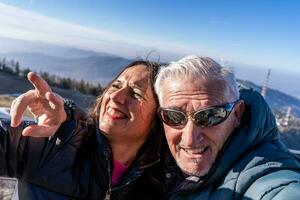 middle aged couple wearing winter clothes taking a selfie over a mountain photo