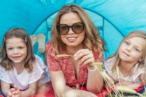 portrait of cute blonde mom with her daughters lying down in a blue camping tent photo