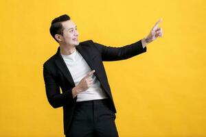 Portrait of happy young Asian man pointing fingers away at copy space isolated on yellow background photo