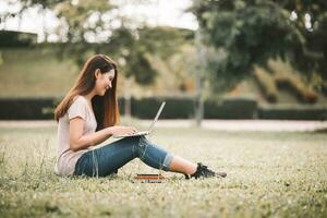 Confident smiling young Asian woman students reading and thinking doing homework with computer laptop on lawn in park. Concept of Education, Campus, teenage. photo