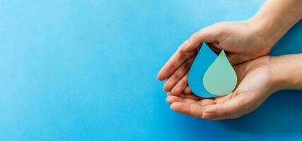 Woman hands holding paper cut water drop on blue background. Clean water and sanitation, saving water. World Water Day. Ecology concept. photo