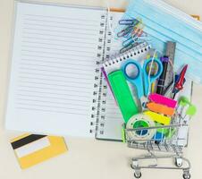 Shopping cart with school supplies, protective mask, alcohol gel. Back to school during a covid-19 pandemic. photo