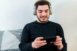 Front view of a happy man with headphones using a mobile phone on line sitting on a couch photo