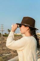 Young woman with brown hat and beige sweater, looking through binoculars, in natural park. photo