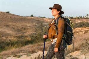 Young woman hiker with leather jacket, hat, backpack, and tracking stick, walks through a field. photo