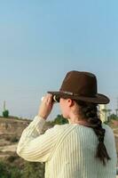 Young woman with brown hat and beige sweater, looking through binoculars, in natural park. photo