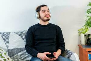 Front view of a happy man with headphones using a mobile phone on line sitting on a couch photo