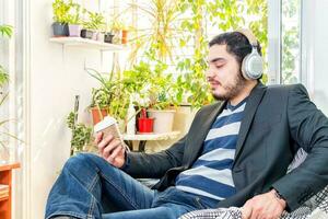 joven barbado hombre escuchando a música con inalámbrico auriculares, participación un café a Vamos en papel taza. foto