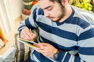 A serious young student is sitting on the couch and writing some plans. He is taking his notes. photo