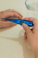 Female hands holding a digital thermometer, tablets in blisters, a glass of water, on desk. Concept of fever, flue or virus. photo