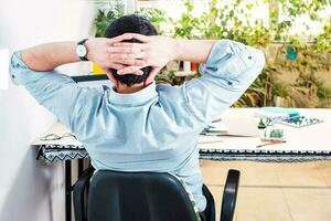 Man relaxing while working remotely from home, looking at the laptop, stretching hands behind his head, with balcony in background. photo