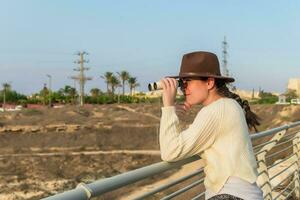 Young woman with brown hat and beige sweater, looking through binoculars, in natural park. photo
