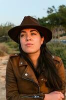 Young woman in hat and brown leather jacket, looking into the distance, in natural park. photo