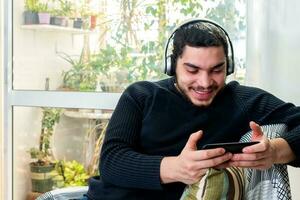 Front view of a happy man with headphones using a mobile phone on line sitting on a couch photo