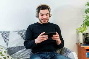 Front view of a happy man with headphones using a mobile phone on line sitting on a couch photo