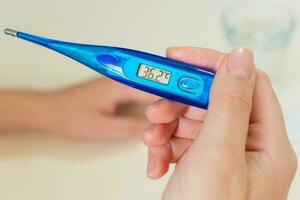 Female hands holding a digital thermometer, tablets in blisters, a glass of water, on desk. Concept of fever, flue or virus. photo