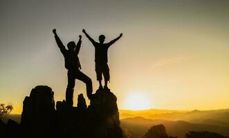 Silhouette of  Hikers climbing up mountain cliff.  Teamwork of two men hiker helping each other on top of mountain climbing team beautiful sunrise landscape. photo