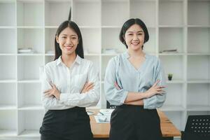 retrato de joven sonriente dos mujer mirando a cámara con cruzado brazos. contento dos niña en pie en creativo oficina. exitoso mujer de negocios en pie en oficina con Copiar espacio. foto