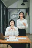 Happy business woman standing competently and smiling in open plan office with blurry collea  gues sitting in office as background, Business concept. photo