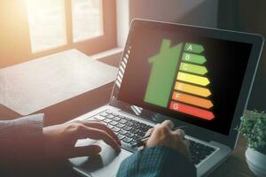 Person hand using laptop computer with house icon energy efficiency scale image on office desk, Concept of ecological and bio energetic house. Energy class. photo