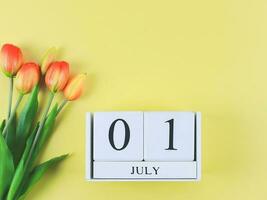 flat lay of wooden calendar with date July 01 on yellow  background with orange and yellow tulips, copy space. photo