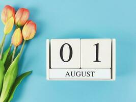 flat lay of wooden calendar with date August 01 on blue  background with orange and yellow tulips, copy space. photo