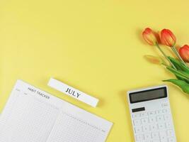 plano diseño de hábito rastreador libro, blanco calculadora, de madera calendario julio y tulipanes en amarillo antecedentes. foto