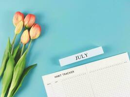 flat lay of habit tracker book with wooden calendar July,   and tulips  on blue background. photo