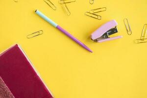 Stationery is on a yellow background. A pen and a stapler are lying next to a pink notebook photo