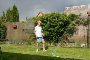 A cute boy is standing on the lawn under drops of water. Running under the sprinkler photo