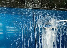 Blue car wash with white soap foam and professional auto care service. Car cleaning service concept. Vehicle cleaning service. Foam wash car detailing. Blue luxury SUV car is covered with white foam. photo