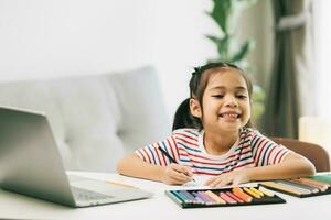 pequeño asiático niña dibujo con color lapices y ordenador portátil a hogar foto