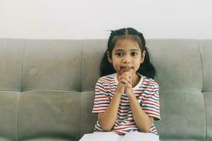 Little asian girl praying for god and reading a book on sofa in living room at home photo