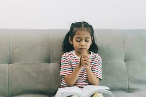 Little asian girl praying for god and reading a book while sitting on sofa at home photo