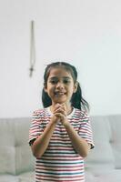 Little asian girl smiling and looking at camera while sitting praying for god on sofa at home photo