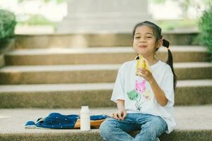 pequeño asiático niña sentado en escalera y teniendo desayuno con Leche y banana. foto