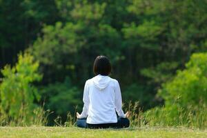 Rear view of woman in hoodie is relaxingly practicing meditation yoga in forest full of grass meadow in summer to attain happiness from inner peace wisdom with morning light for healthy mind and soul photo