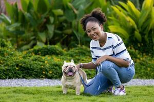 African American woman is playing with her french bulldog puppy while walking at the dog park at grass lawn after having morning exercise during summer photo