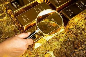 a hand with a magnifying glass checks the gold bar on a pile of gold coins photo
