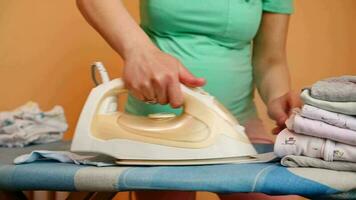 Selective focus on hand of a pregnant woman holding electric steam iron, ironing laundered clean newborn baby clothes, folding them on ironing board, preparing bag for the maternity hospital. Closeup video