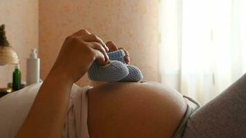 de cerca. barriga de un embarazada mujer esperando un bebé poniendo bebé botines en su grande barriga, disfrutando bebé movimientos, relajante acostado en el cama a hogar dormitorio. el embarazo. maternidad. contento maternidad video