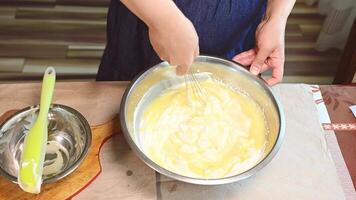 Visão a partir de acima do uma dona de casa em pé às cozinha bancada, usando bata, mexendo líquido ingredientes com açoitado creme dentro uma metal tigela, preparando gelificado mousse para uma bolo às lar. confeitaria video