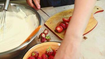 Close-up chef's hands slicing strawberries in halves on a wooden board, for decorating a mousse dessert Fraisier. Whipped cream in metal bowl on the kitchen worktop. Confectionery. Bakery. Culinary video